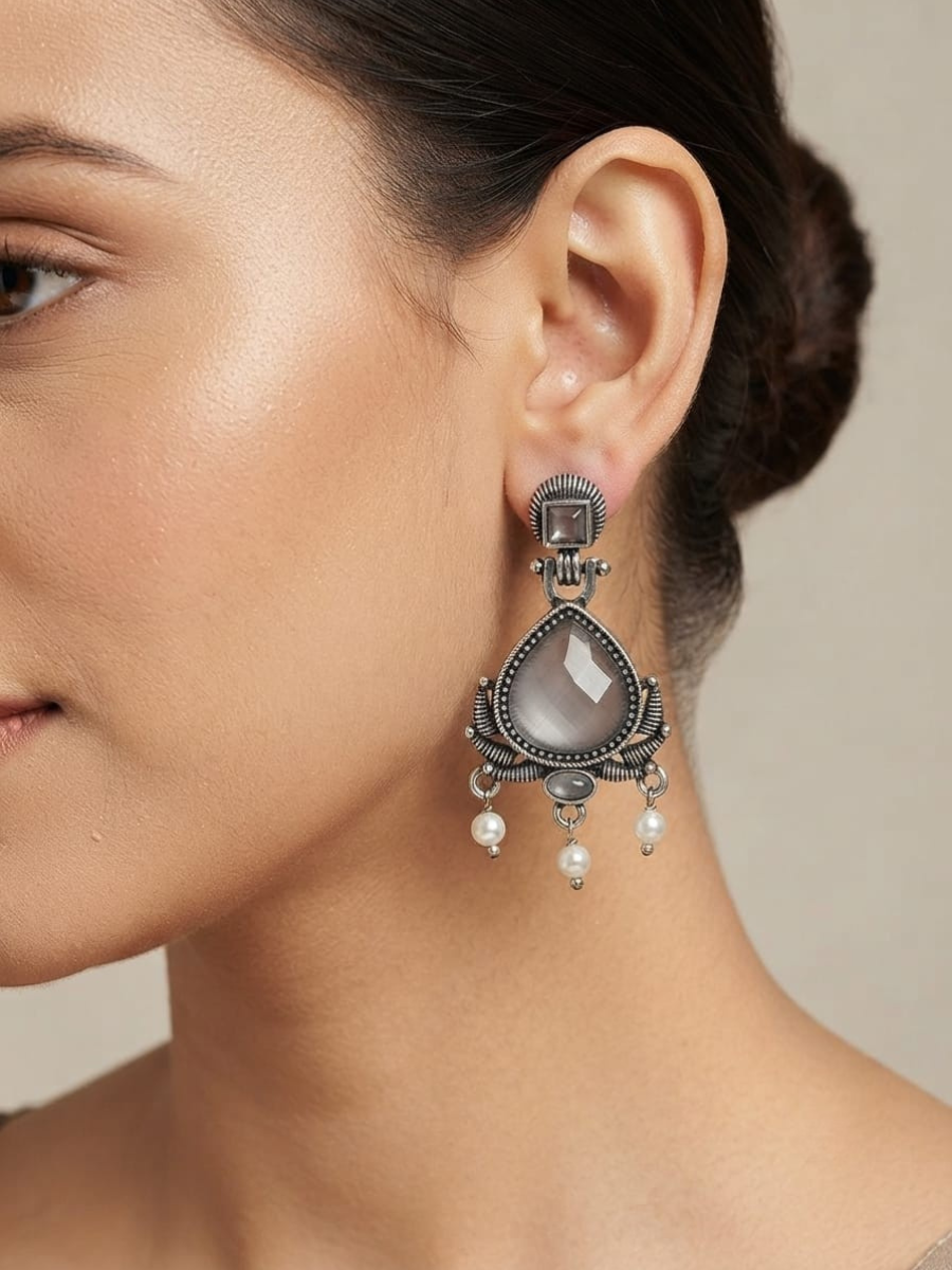 A close-up of a woman's ear wearing a large silver-toned dangle earring with a teardrop-shaped pale stone and small pearl-like drops.