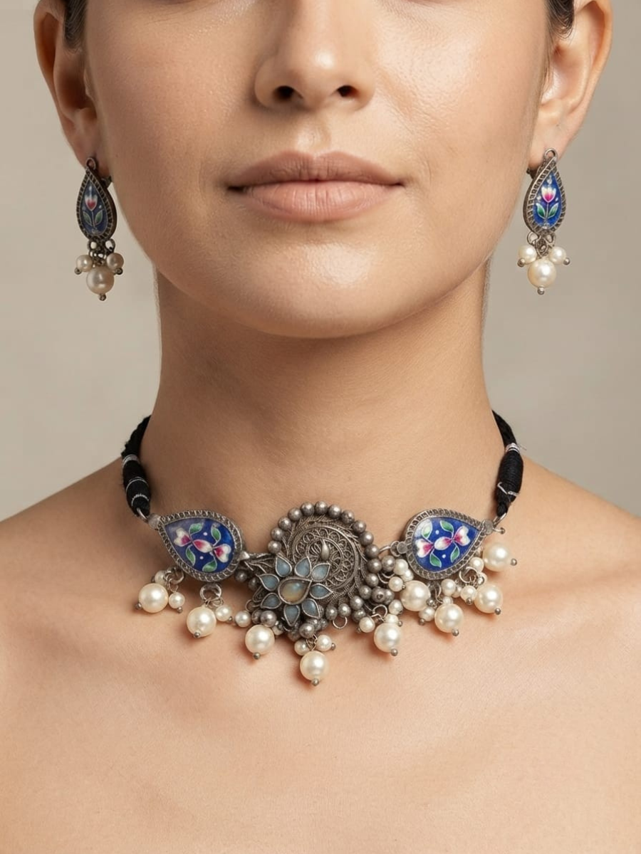 A close-up of a model wearing a silver-toned statement necklace with blue, pink, and white enamel details, pearl-like beads, and a black cord, along with matching drop earrings.