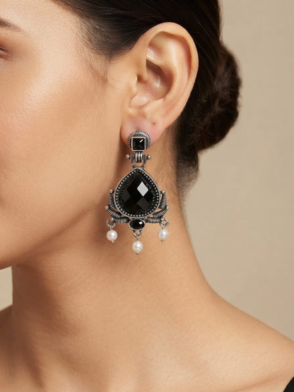 A close-up of a model wearing a dramatic black teardrop dangle earring with silver filigree detailing and small pearl drops.