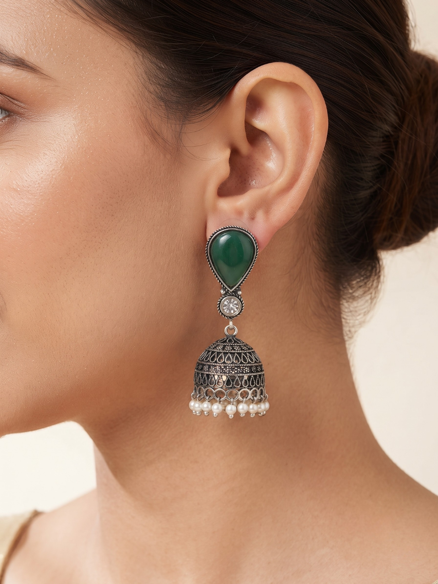 A close-up of a model wearing a traditional oxidized silver pearl drop earrings The earring features a teardrop-shaped green stone set in silver, connected to a silver filigree bell with small white pearl drops at the bottom.