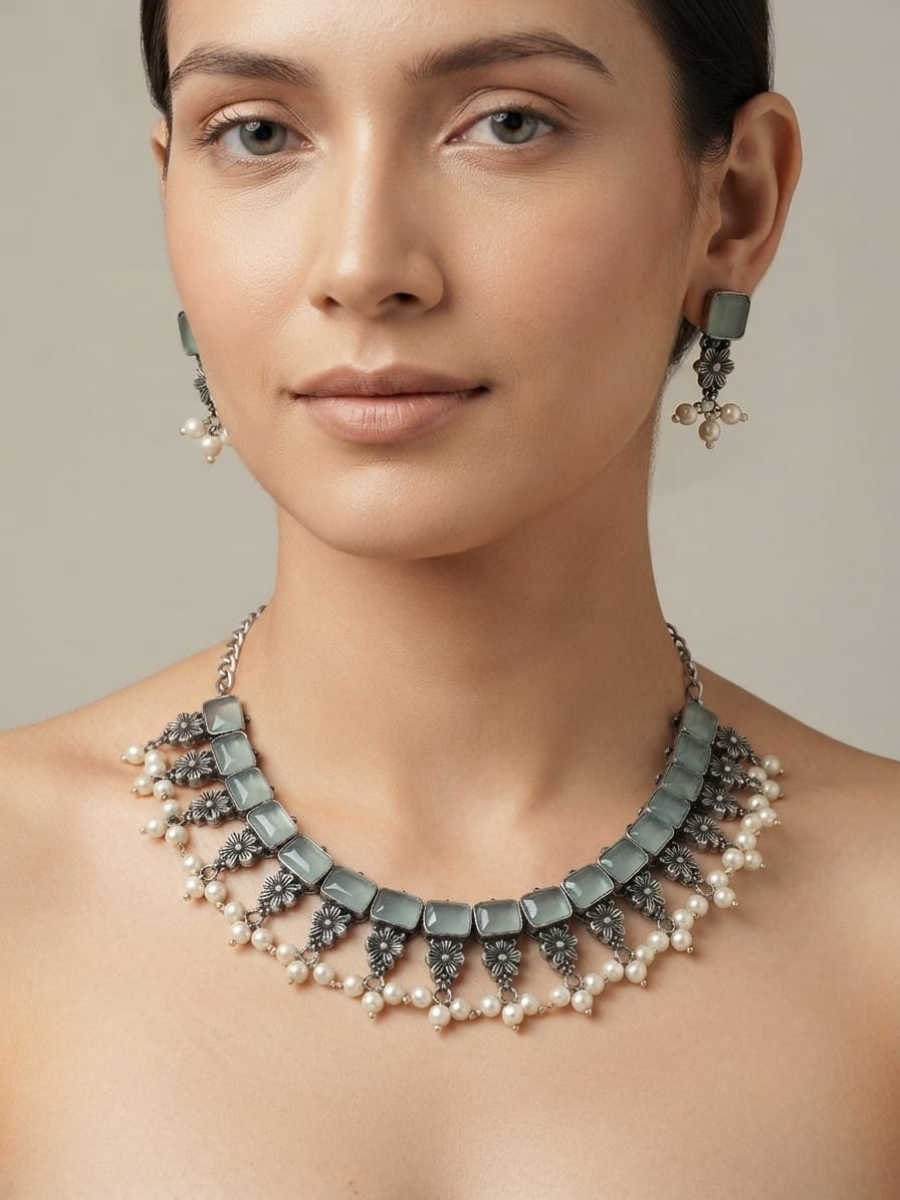 A close-up of a model wearing a statement necklace and matching drop earrings. The jewelry features rectangular light mint-green stones, dark gray filigree details, and rows of small white pearls
