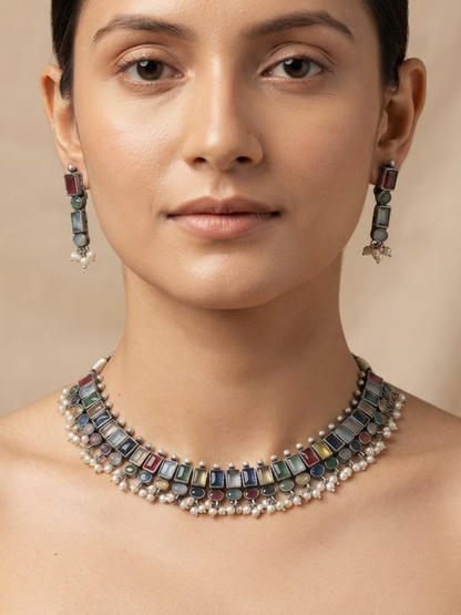 A close-up of a model wearing a multi-strand statement necklace and matching drop earrings. The jewelry features rectangular multicolored gemstone accents in shades of blue, green, red, and gold, accented by rows of small white pearls