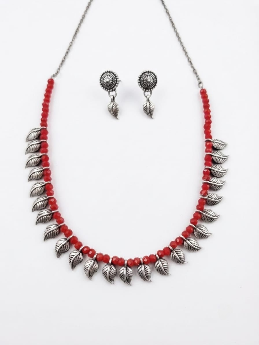 A jewelry set featuring a silver-toned necklace and matching earrings. The necklace is composed of small red beads, silver-toned leaf-shaped charms, and a silver-toned chain.