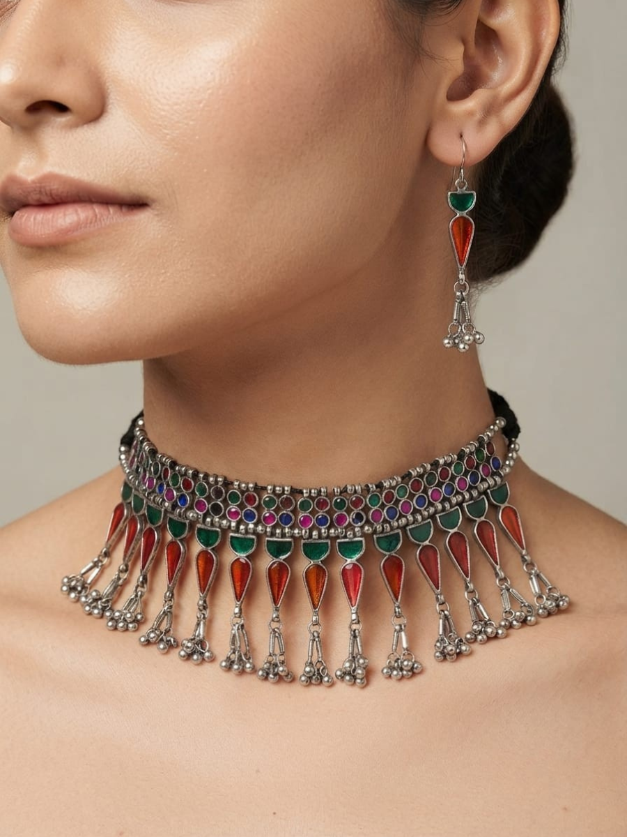 A close-up of a woman wearing a silver-toned choker necklace with dangling red and green enamel teardrop charms, paired with a matching drop earring.