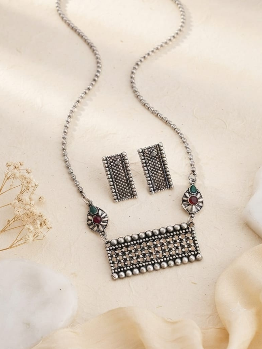 A silver-toned traditional jewelry set featuring a rectangular pendant necklace with a beaded border, matching rectangular earrings, and small decorative elements with red and green stones
